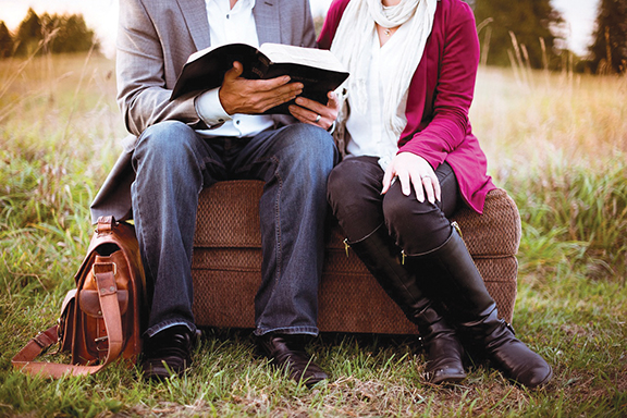 Couple seated on sofa in field, reading a book, wearing casual fall attire with a brown leather bag beside them.
