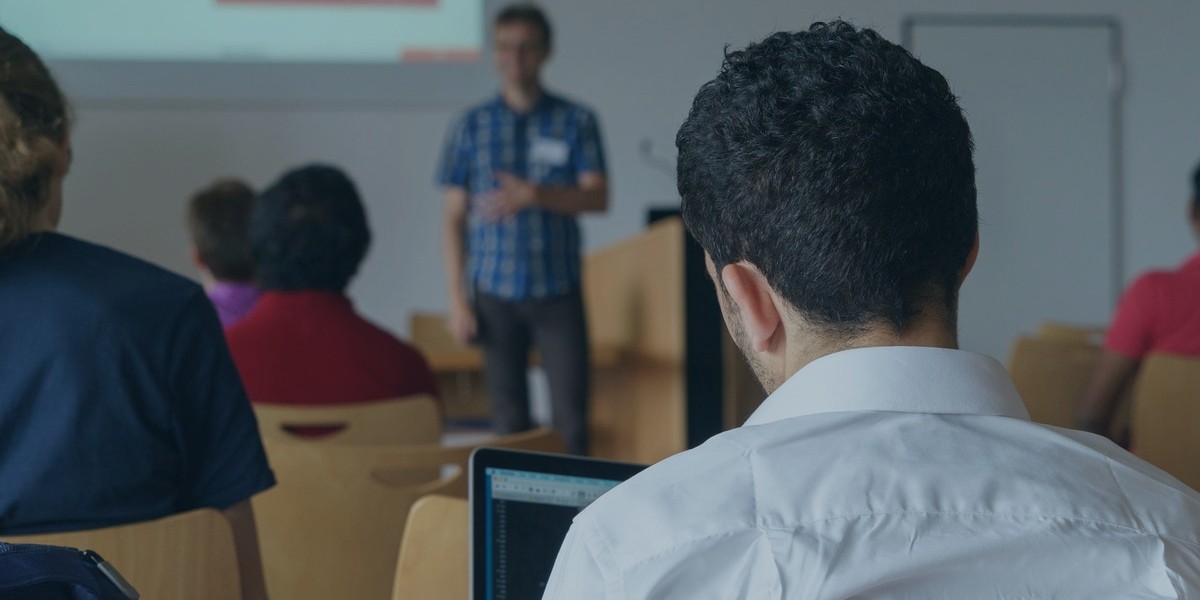 Student using laptop in classroom with lecturer speaking at the front, presentation in progress.