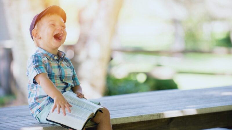 A joyful child in a plaid shirt and cap laughs while holding an open book outdoors on a sunny day.