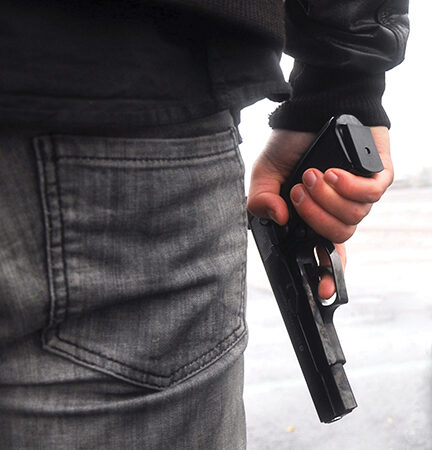 Person holding a handgun behind their back, wearing jeans and a jacket, standing outdoors.
