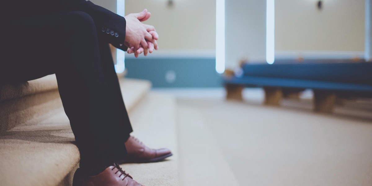 Man in suit sitting on steps with hands clasped, empty church interior in background.