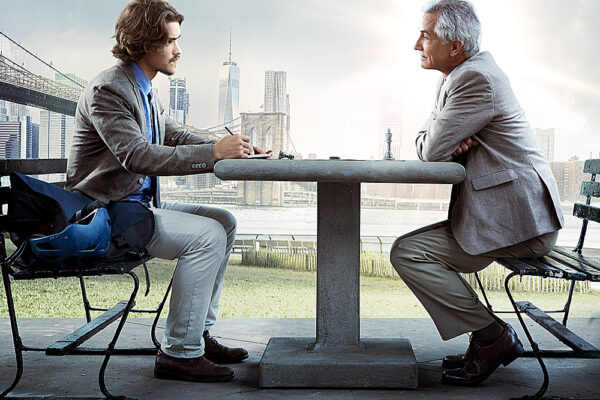 Two men conversing at a park table with a cityscape background, sunlight shining through clouds.