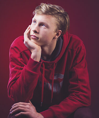 Young man in red hoodie, hand on chin, gazes thoughtfully upward against a red background.