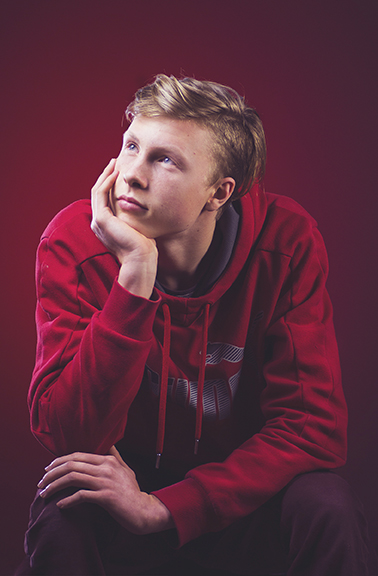 Young man in red hoodie, hand on chin, gazes thoughtfully upward against a red background.