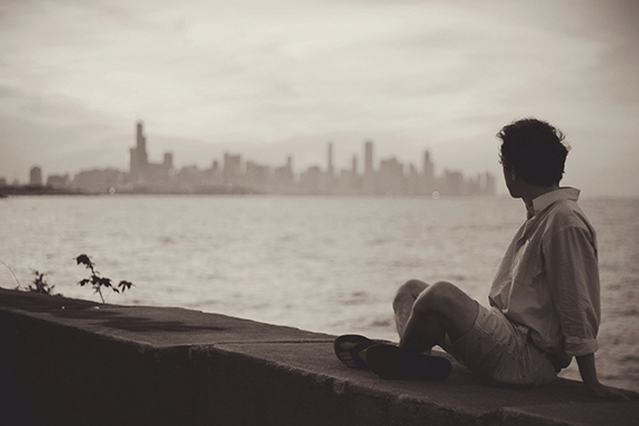 Person sitting on a wall gazing at an urban skyline across the water in sepia tones.