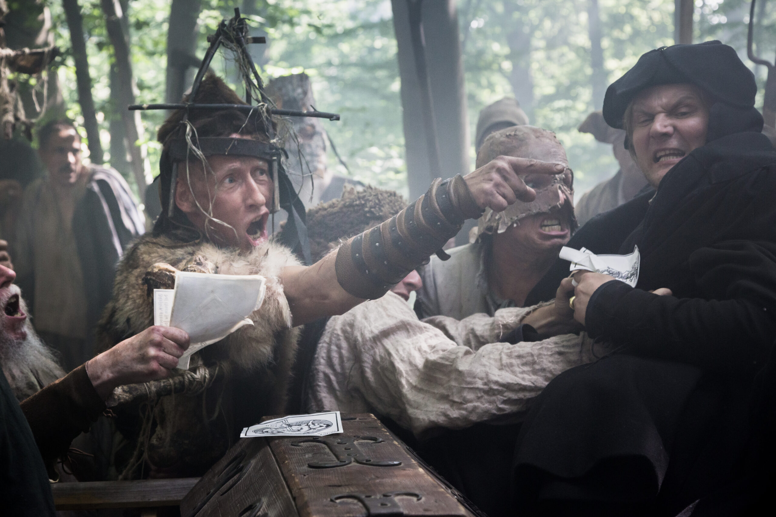 Medieval men in intense confrontation, one in fur and metal headgear, others gesturing and holding papers in a forest setting.