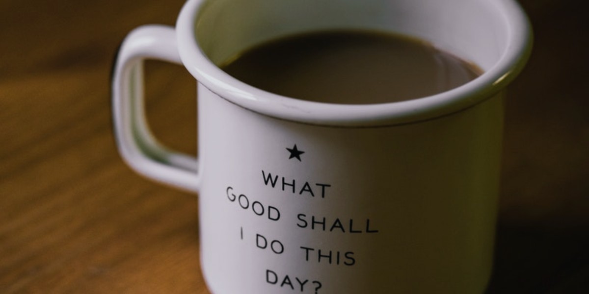 Ceramic mug with motivational quote What good shall I do this day? filled with coffee on wooden table.