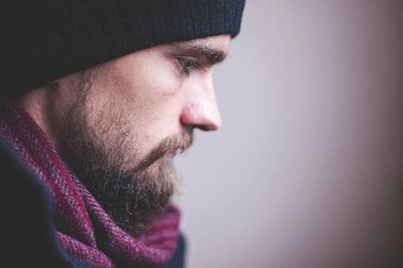 Man in a beanie and scarf gazes thoughtfully downward, side profile, with a neutral background.