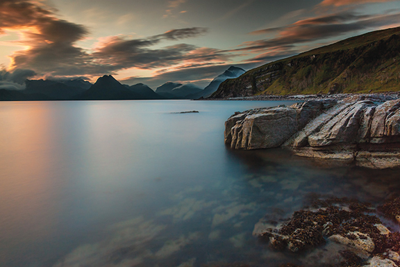 Tranquil coastal scene with rocky shoreline at sunset, featuring calm waters and a dramatic sky.