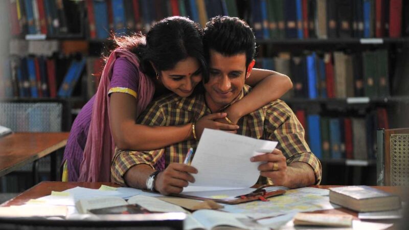 Couple reading and embracing in a library, surrounded by books and papers, with shelves in the background.