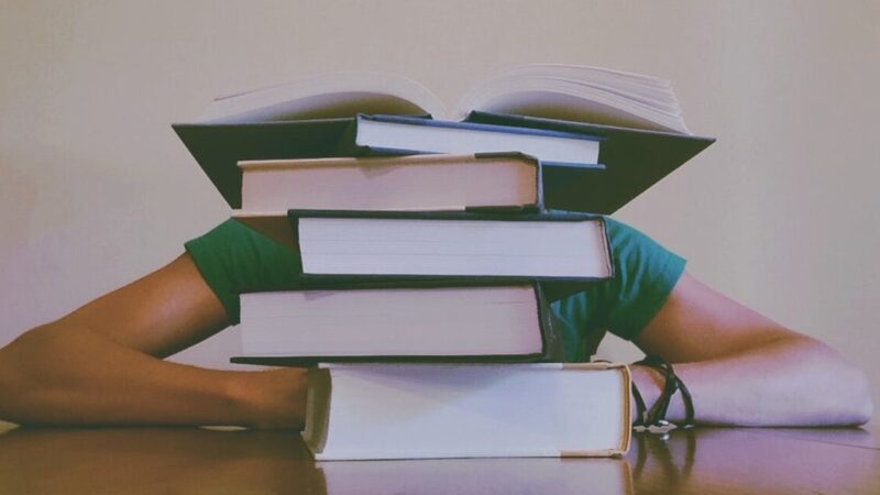 Person overwhelmed by a stack of books, highlighting a challenging study session.