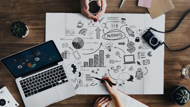 Creative business strategy planning on paper with laptop, charts, and doodles, surrounded by hands and coffee on a desk.