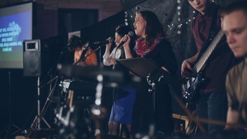 Live band performing on stage with singers and musicians, dimly lit, screen in background showing lyrics.