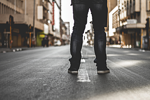 Person standing on an empty urban street, wearing jeans and sneakers, with buildings in the background.
