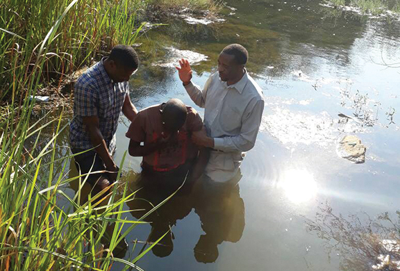 Three men participating in an outdoor baptism ceremony in a natural water setting surrounded by reeds.