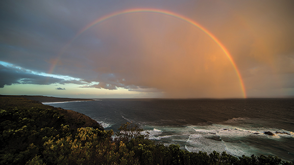 Rainbow over ocean at sunset with dramatic sky and coastal view.