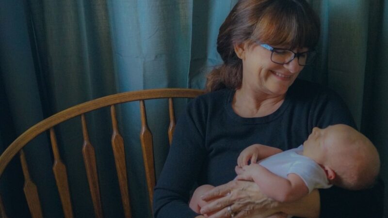 Smiling woman wearing glasses holding a baby in her arms, sitting on a wooden bench with blue curtains in the background.