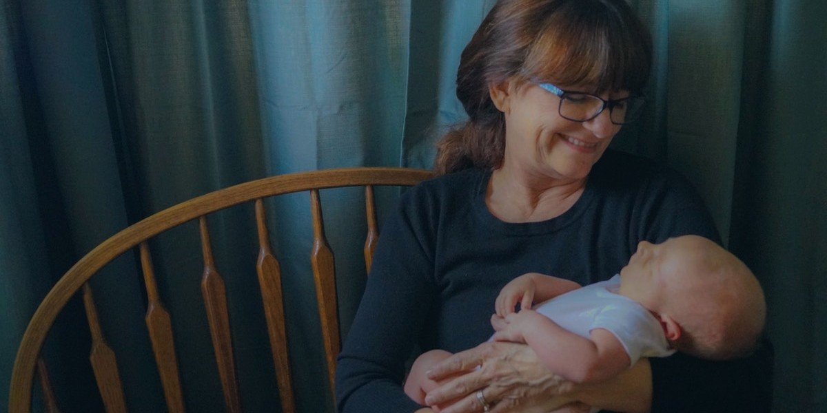 Smiling woman wearing glasses holding a baby in her arms, sitting on a wooden bench with blue curtains in the background.