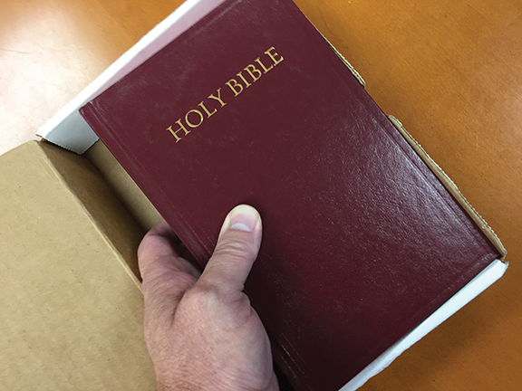 Person holding a red Holy Bible being taken out of a cardboard box on a wooden table.