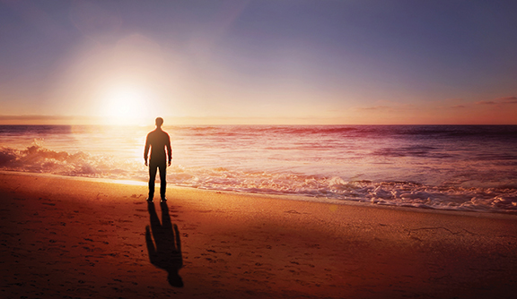 Person standing on a beach at sunset, gazing at the ocean, with sun casting a warm glow on the water and sand.