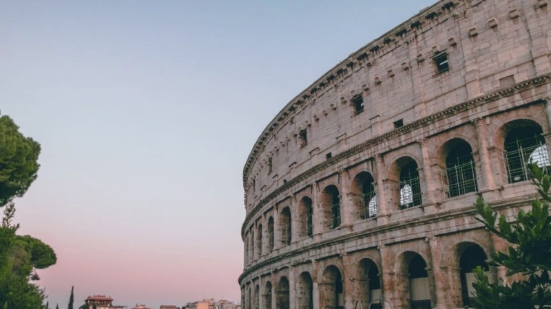 Colosseum at sunset with pink sky, Rome's iconic ancient amphitheater, partially obscured by trees.