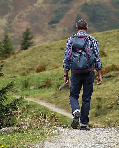 Man hiking on a mountain trail with a backpack, surrounded by lush greenery and trees.