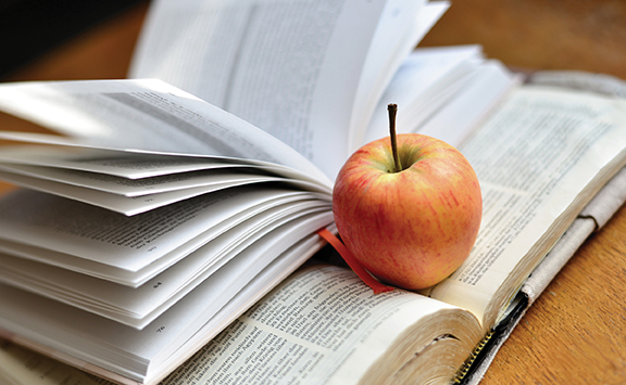 Open book with pages fanned and a red apple on top, symbolizing knowledge and education on a wooden table.