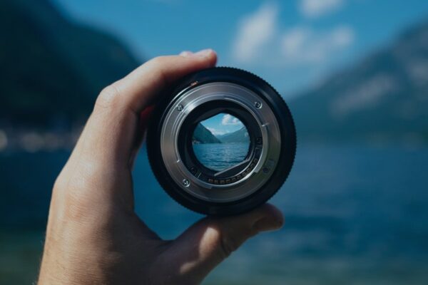 Hand holding a camera lens, focusing on a scenic lake view with mountains in the background.