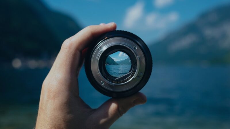 Hand holding a camera lens, focusing on a scenic lake view with mountains in the background.