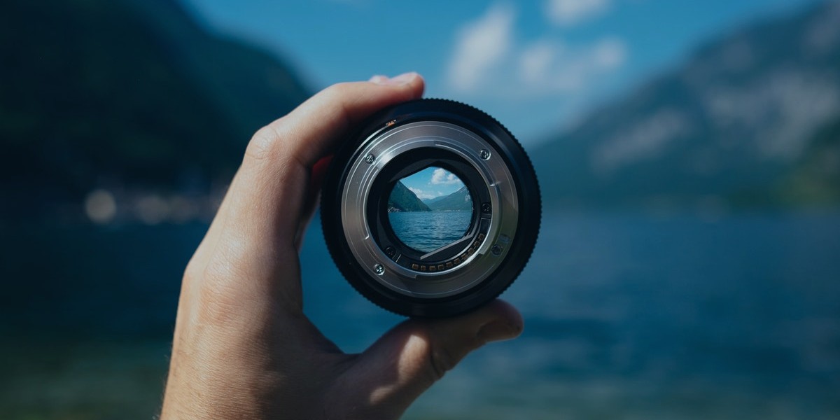 Hand holding a camera lens, focusing on a scenic lake view with mountains in the background.