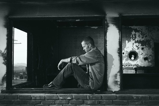 Man sitting thoughtfully in a window frame of a worn building, gazing into the distance in black and white.