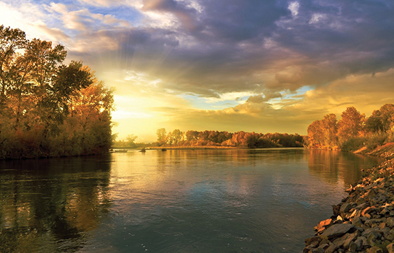 Sunset over a serene river with trees reflecting in the water under a dramatic sky.