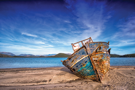 Old, weathered boat on sandy shore with blue ocean and clear sky in the background.