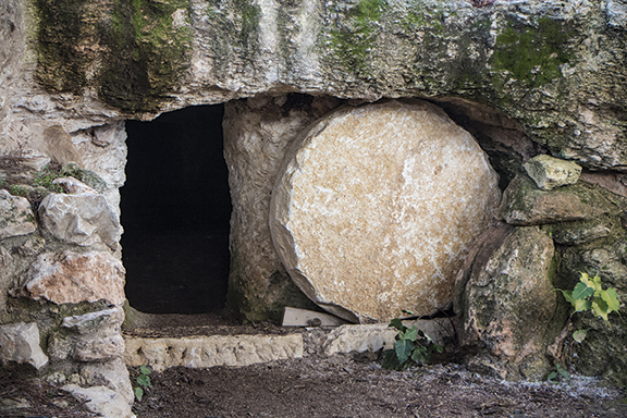 Ancient stone tomb with a large circular rock partially blocking the entrance, surrounded by mossy stones.