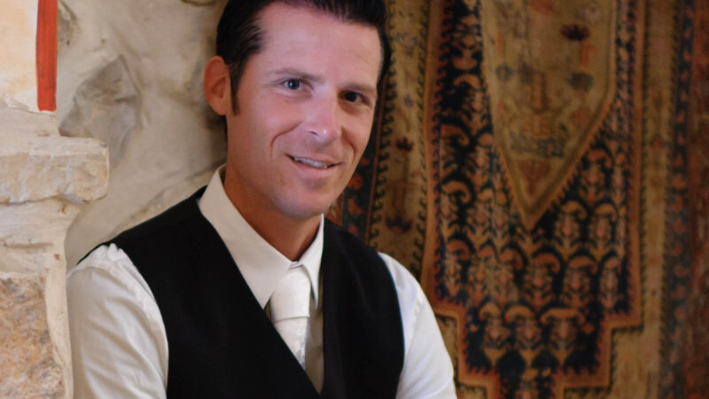 Man in formal attire sitting next to a stone wall and ornate rug, smiling.