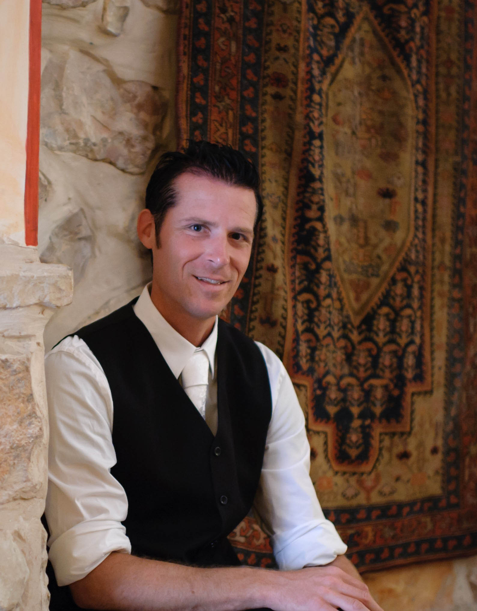 Man in formal attire sitting next to a stone wall and ornate rug, smiling.