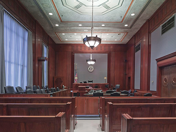 Wood-paneled courtroom interior with empty benches, judge's bench, and large overhead lights.