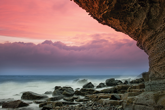 Dramatic coastal cliff at sunset, with pink sky over ocean rocks and waves.