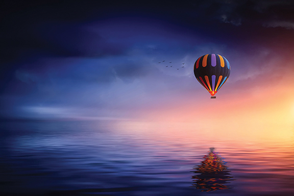 Hot air balloon floating over calm ocean at sunrise, reflecting vibrant colors in water.