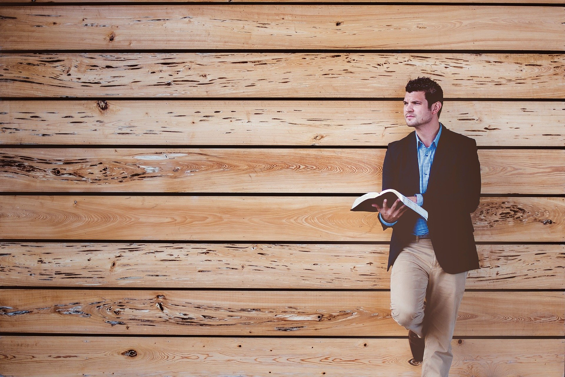 Man in casual attire reading against wood wall.