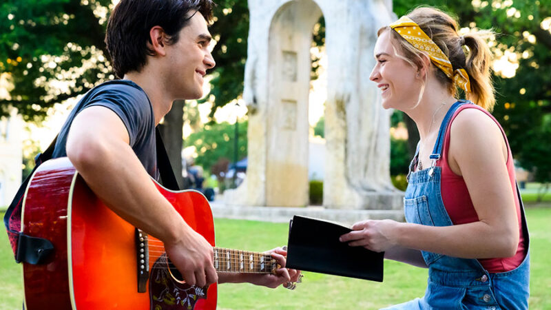 Young couple enjoying an outdoor guitar session in a park, woman holds notebook and smiles at guitarist.