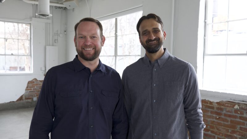 Two smiling men in a bright office setting with windows and exposed brick walls.