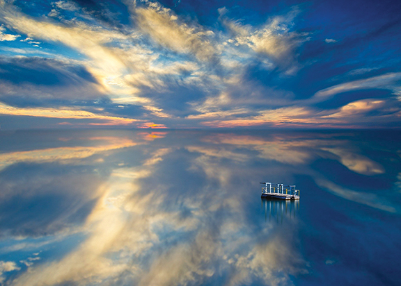 Surreal sunset sky mirrored on a calm lake with a small boat, creating a stunning natural reflection.