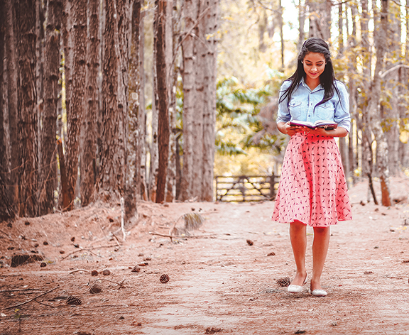 Woman reading in a forest, wearing a denim jacket and pink skirt, surrounded by tall trees and autumn leaves.