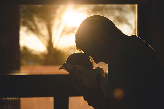 Father holding newborn against sunset backdrop, warm light streaming through window.