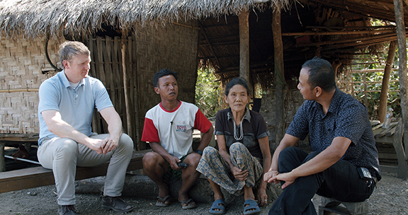 Four people having a discussion outside a traditional thatched hut.