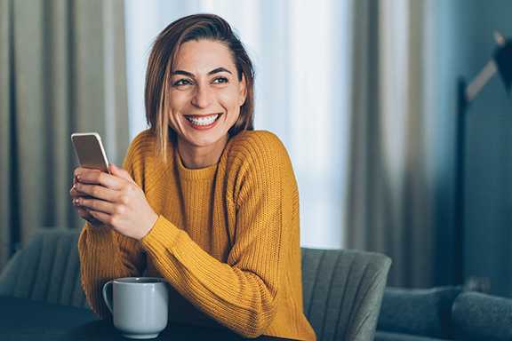 Smiling woman in yellow sweater holding smartphone and coffee mug indoors, feeling happy and relaxed.