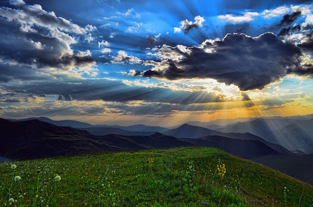 Sun rays breaking through clouds over a scenic mountain landscape with a grassy field in the foreground.