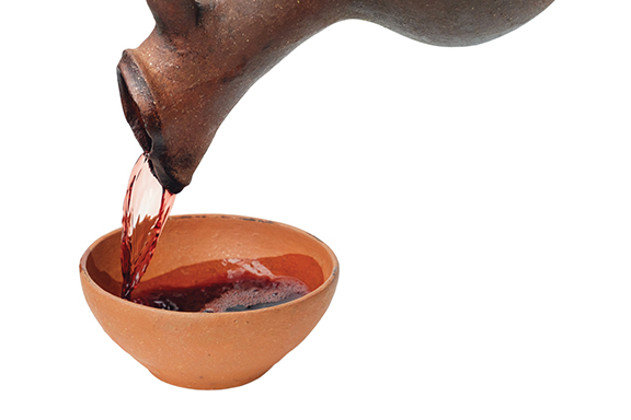 Earthenware jug pouring red liquid into a clay bowl on a white background.