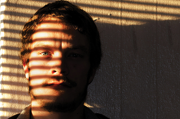 Man's face partially illuminated by sunlight filtering through window blinds, casting striped shadows across his features.